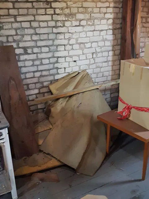 Dachboden vor der Entrümpelung: Klinkerwand, abgedeckte Möbel, Holzplatten und ein Tisch stehen dicht an dicht